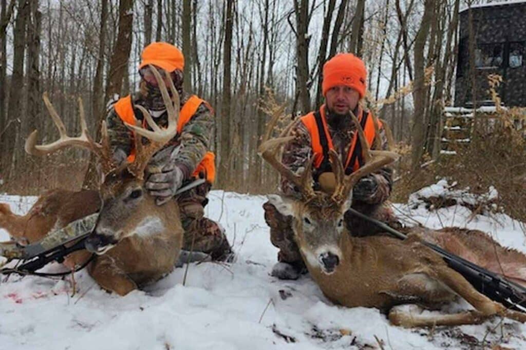 Two hunters in camouflage and orange vests sit in snowy woods, each posing with a rifle and a harvested buck with antlers.