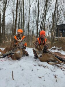 Two hunters in camouflage and orange vests pose in the snow with two large deer they have hunted; bare trees and a dark cabin are visible in the background.