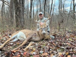 A man in camouflage clothing kneels in a forest next to a large deer with antlers lying on the ground among fallen leaves.