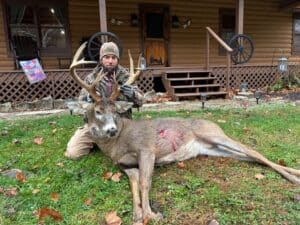 A person kneels behind a large deer with antlers on a lawn in front of a wooden house.