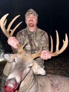 A person in camouflage clothing holds the antlers of a large deer with visible blood on its nose, photographed at night.