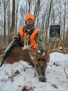 Person in camouflage and orange hunting gear poses with a rifle beside a large buck deer in a snowy forest, with a hunting blind visible in the background.